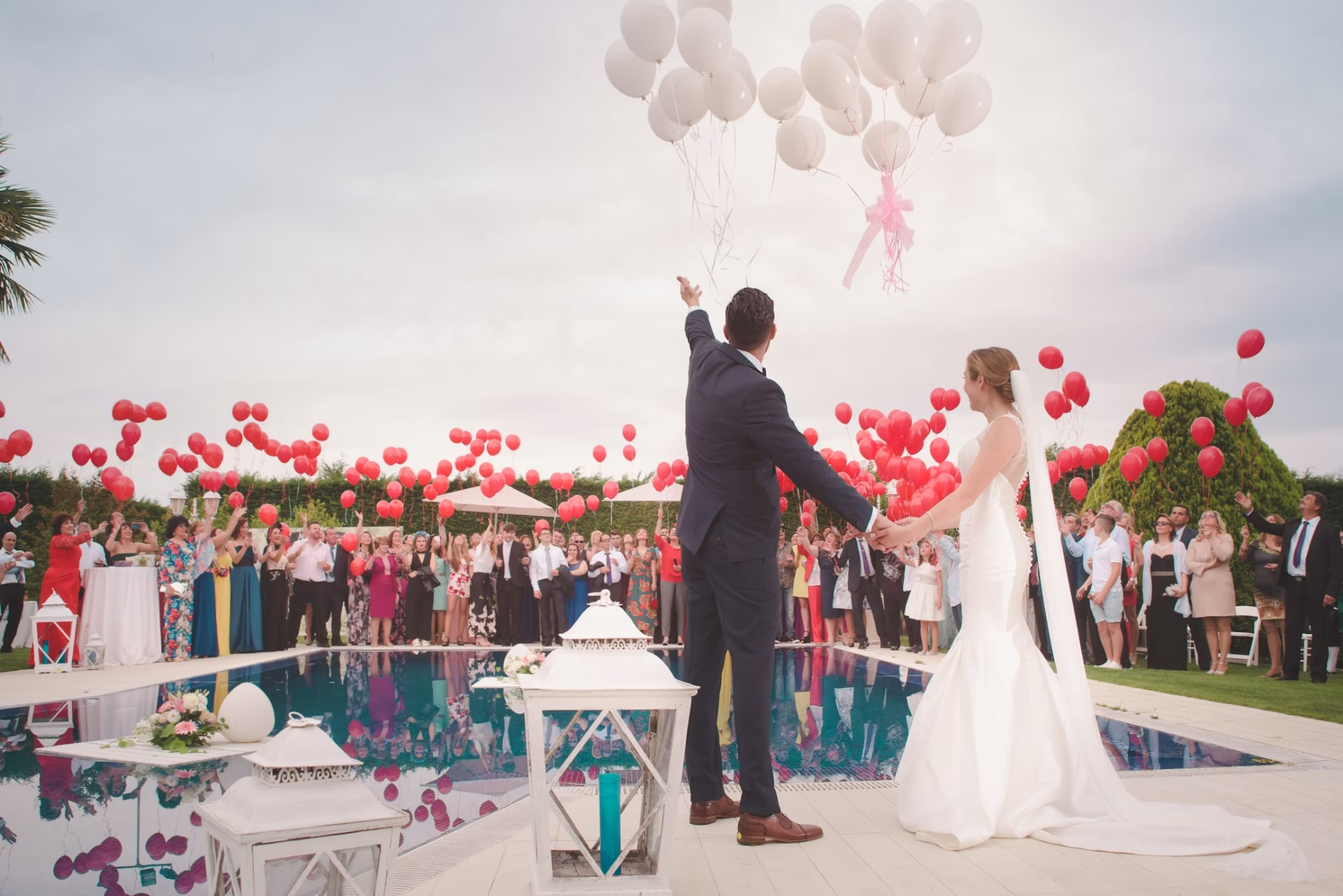 Bride and groom releasing balloons with guests cheering at a poolside wedding celebration